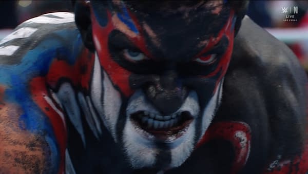 A wrestler with intricate face paint featuring red, black, and blue patterns is shown in an intense expression. The focus is on his fierce gaze and painted features, creating a striking visual impact.