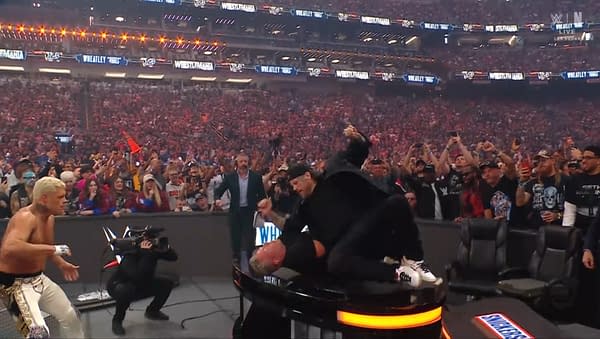A wrestling match scene captures an intense moment where a man in black is attacking another man on a round table. In the background, a large crowd of fans is cheering in an arena filled with excitement.