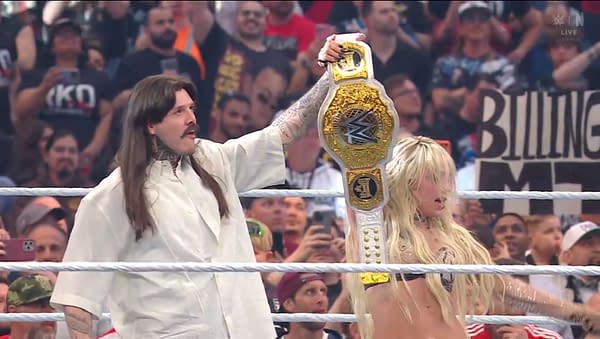 A wrestler holds a championship belt high above their head, celebrating a victory in the ring. Another wrestler stands beside them, both surrounded by a cheering crowd.