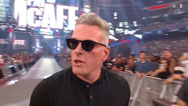 A man with short, silver hair wearing sunglasses walks past a crowd at a wrestling event, with bright lights and a large screen in the background displaying 'The McAfee'.