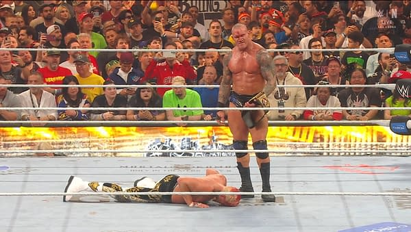 A muscular wrestler stands triumphantly in a wrestling ring, holding a championship belt, while another wrestler lies defeated on the mat. Fans fill the background, capturing the moment on their phones.