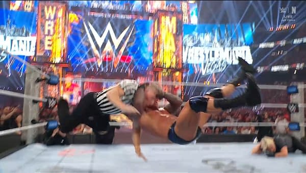 A dynamic wrestling scene showing a referee executing a move on a wrestler, with bright lights and a large WWE WrestleMania sign in the background. The atmosphere is energetic and intense.