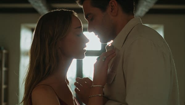 A softly lit romantic close-up of a couple leaning in forehead to forehead, with the woman touching the man's shirt in a sunlit room.