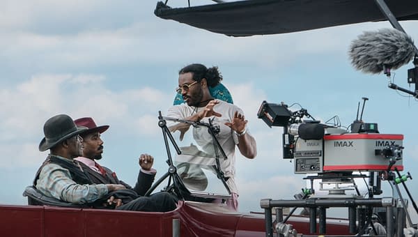 A scene from a film set featuring Ryan Coogler engaging with two actors seated in a vintage car, while an IMAX camera is positioned nearby. The sky is partly cloudy, and the atmosphere suggests a behind-the-scenes look at filmmaking.