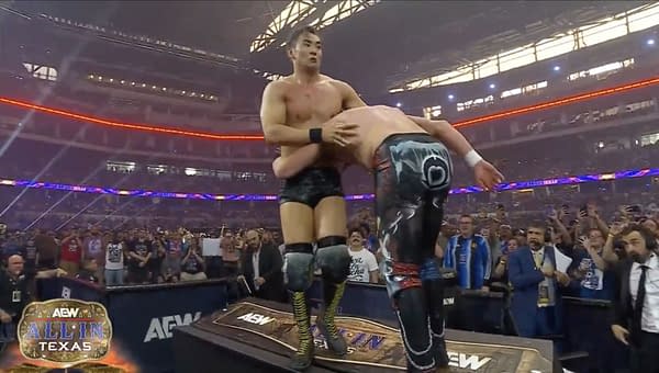 Kazuchika Okada and Kenny Omega engage in a grappling contest on the announce desk during AEW All In Texas, with an energetic crowd visible in the background.