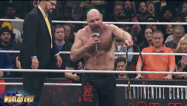 Jon Moxley delivers a passionate promo in the wrestling ring at AEW World's End, holding a championship belt and addressing fans and wrestlers. A commentator stands nearby while the enthusiastic audience is visible in the background.