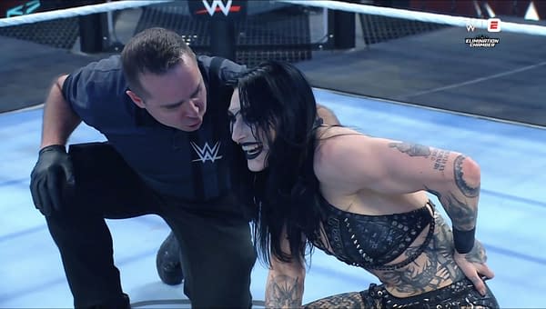 Rhea Ripley, wearing a black outfit with tattoos visible, celebrates her victory in the Elimination Chamber, smiling and engaging with a referee.