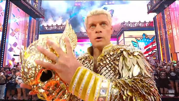 A wrestler with blonde hair and a golden, intricately designed outfit is holding an ornate gold mask while looking intensely at the camera. The background has colorful lights and a cheering crowd.