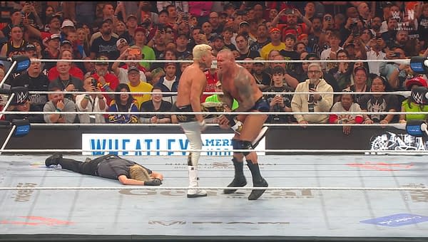 Cody Rhodes is in a wrestling ring, facing an opponent while a referee is down on the mat. The audience is engaged, with many fans holding up phones to capture the moment. Spectators include a mixed crowd with visible excitement during the match.