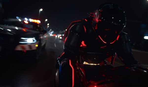 A scene from Disney's live-action film "TRON: ARES," featuring a futuristic character in a sleek, illuminated black and red suit riding a motorcycle, closely pursued by a police car with flashing lights, set against a nighttime urban backdrop.