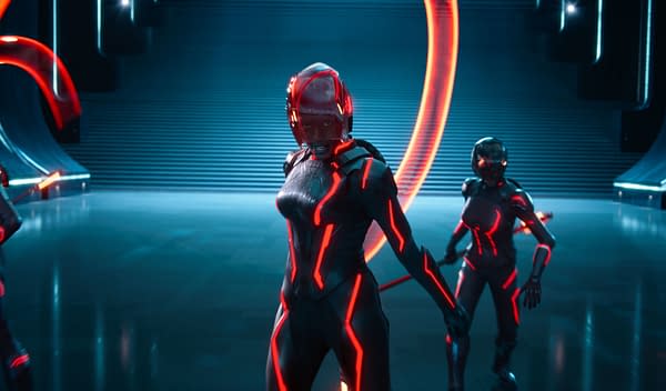 Jodie Turner-Smith as Athena stands confidently in a futuristic setting, wearing a sleek black suit with glowing red lines. Two other characters in similar outfits accompany her, set against a backdrop of illuminated structures and a staircase.