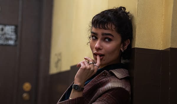 A young woman with dark curly hair, wearing a plaid coat, leans against a wall with a playful expression, finger touching her lips. The background features a brown door and a brightly colored wall.