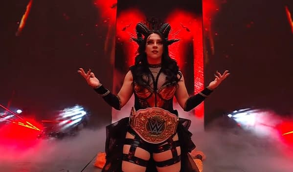 A female wrestler poses confidently with the WWE Championship belt around her waist, set against a dramatic red and black background with fog and lighting effects. She has a striking outfit featuring a fishnet top and an elaborate headdress.