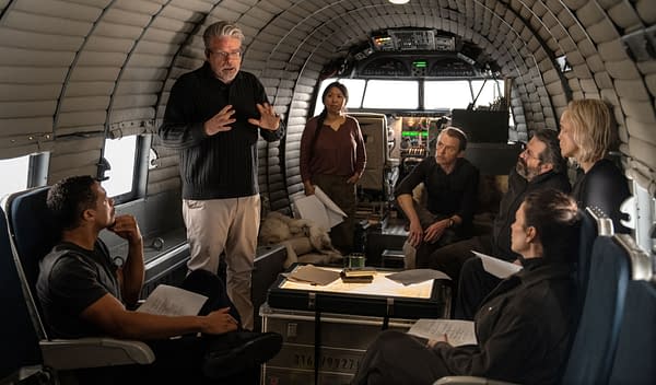 A scene from 'Mission: Impossible - The Final Reckoning' showing a group of six involved in a discussion on a set designed like an aircraft interior. The director is gesturing while actors focus on the conversation, with scripts and a table of documents visible.