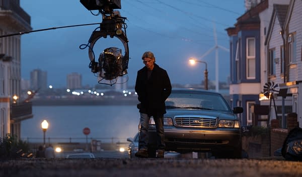 A moody film-set scene at dusk shows a man standing on a street beside a parked car while a camera rig hangs overhead, suggesting Clayface filming.