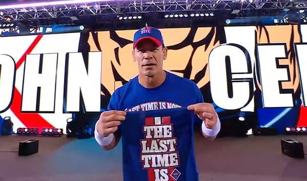 John Cena walks to the wrestling ring wearing a blue shirt that says "THE LAST TIME IS" while displaying a small item in his hand. The background features large graphics of his name.
