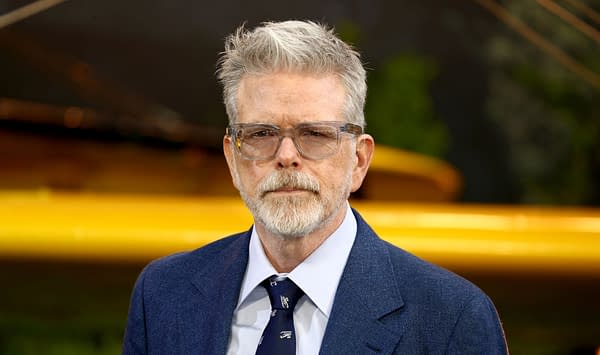 American director Christopher McQuarrie is wearing glasses and a blue suit, with a serious expression, at an event in London. The background includes hints of green foliage and a yellow element.