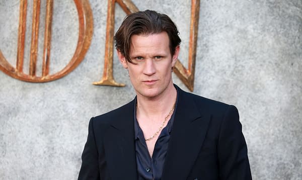 Actor Matt Smith poses confidently, wearing a dark suit and open shirt, against a textured background featuring the word 'LONDON' in gold letters.