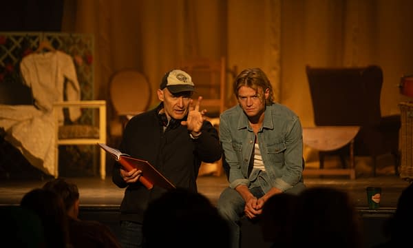 A director and actor sit on a stage during a Clayface production meeting, with the director gesturing while holding a script. Warm backstage lighting and costume racks set the rehearsal scene.