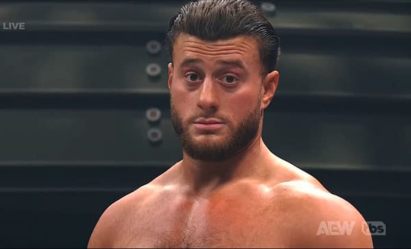 A professional wrestler shows a serious expression in response to a loud crowd chant during a wrestling match. The background features blurred steel structures typical of a wrestling arena.