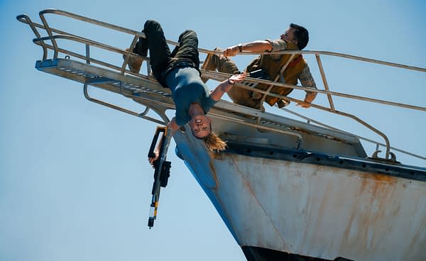 A scene from 'Jurassic World Rebirth' featuring Zora Bennett, played by Scarlett Johansson, hanging upside down from a metal railing, holding a futuristic weapon, while Dr. Henry Loomis, played by Jonathan Bailey, assists her from above. The background shows a clear blue sky.