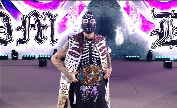 Dominik Mysterio enters the ring at WWE SummerSlam, wearing a skeletal-themed robe and a mask, holding a championship belt. Bright stage lights illuminate the backdrop while he poses confidently for the audience.
