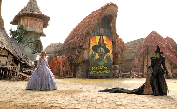 A scene from 'Wicked: For Good' featuring Glinda, dressed in a shimmering gown with a wand, and Elphaba, in a dark, flowing outfit with a broom, standing in a whimsical village. A colorful mural warns 'Beware the Wicked Witch' in the background.