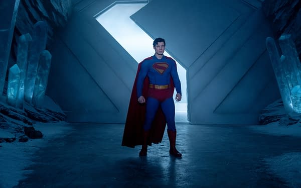 Superman stands confidently in a futuristic ice cave, wearing his iconic blue suit with a red cape. Crystalline structures are visible in the background against a blue-lit atmosphere.
