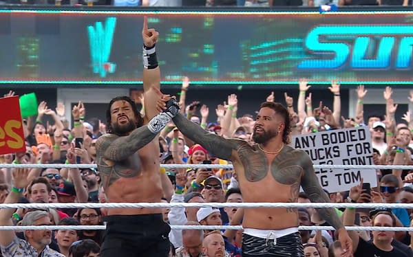 Roman Reigns and Jey Uso celebrate their victory in a wrestling ring at WWE SummerSlam, raising each other's hands amidst a cheering crowd. Signs and excited fans fill the background.