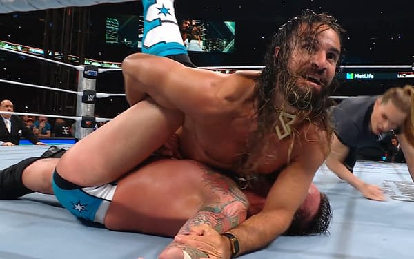 A wrestler with long hair and a beard, Seth Rollins, is pinning another wrestler, CM Punk, down in a wrestling ring. Rollins appears focused and engaged in the match, while the referee is also present in the scene.
