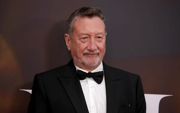 A half-length portrait of a middle-aged man with a greying beard, wearing a black tuxedo, and smiling while attending a film event. The background features a blurred dark gradient, enhancing the focus on his formal attire and expression.