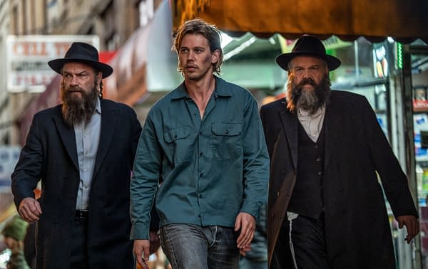 Three men walk through a city street, two in dark clothing and hats, and one in a blue shirt and jeans, suggesting an intense narrative. The setting conveys a sense of urgency and tension.