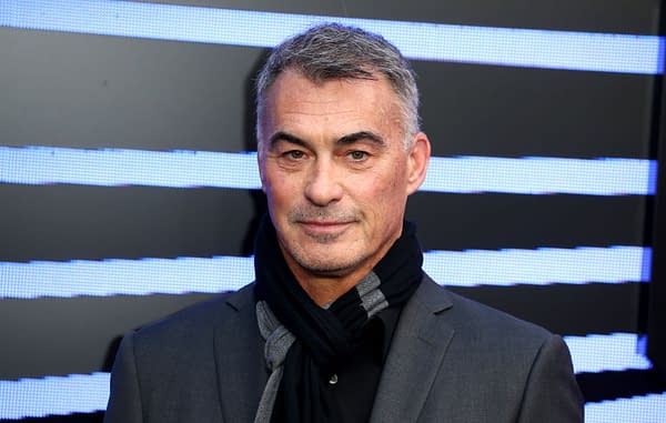 A portrait of Chad Stahelski, an American filmmaker and stuntman, wearing a black suit and scarf, standing in front of a backdrop with blue light stripes.