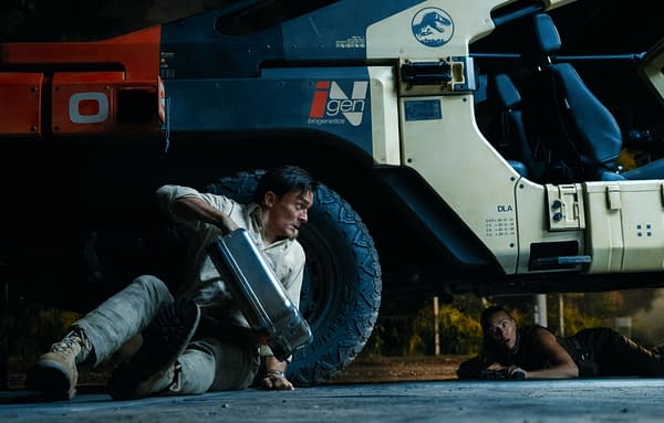 Scene from 'Jurassic World Rebirth' featuring Rupert Friend as Martin Krebs and Scarlett Johansson as Zora Bennett, hiding beneath a large vehicle with a Jurassic Park logo, suggesting an intense covert operation.