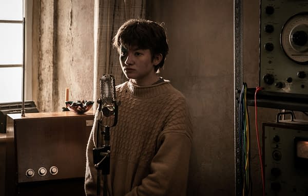 A young person stands near a vintage microphone, wearing a cozy, oversized sweater. The background features an old radio and various audio equipment, creating a nostalgic atmosphere.