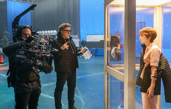 Behind-the-scenes image from 'Black Phone 2' featuring a director guiding the filming process with a camera, while a young actress stands in a glass phone booth next to an old-fashioned payphone.