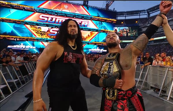 A victorious Solo Sikoa celebrates in the ring after winning a cage match at WWE SummerSlam, holding a championship belt. A teammate stands beside him, both amid a cheering crowd and a colorful arena backdrop.