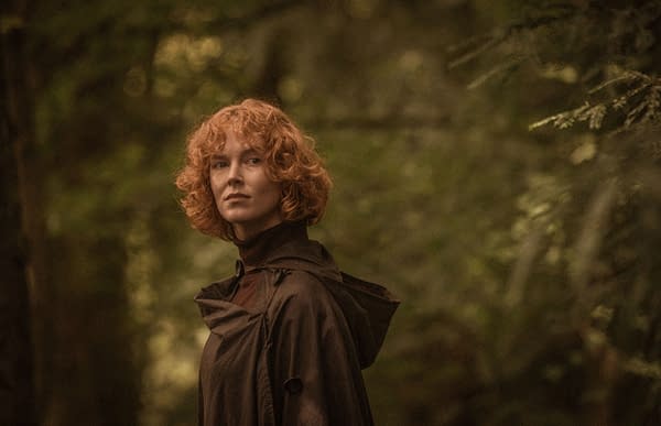 A woman with curly red hair stands in a forest, wearing a dark cloak, surrounded by softly blurred greenery. The warm tones create a serene and contemplative atmosphere.