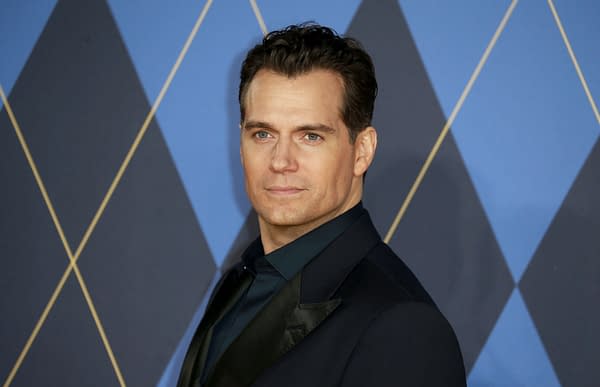 A headshot of Henry Cavill, a British actor, taken during an event in London. He is wearing a dark suit and looking confidently towards the camera against a geometric blue background.