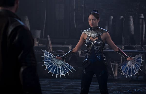 A female character from Mortal Kombat 2 stands ready for battle, wearing a metallic outfit with intricate designs and holding a pair of bladed fans. The dark and atmospheric setting adds intensity to the scene.