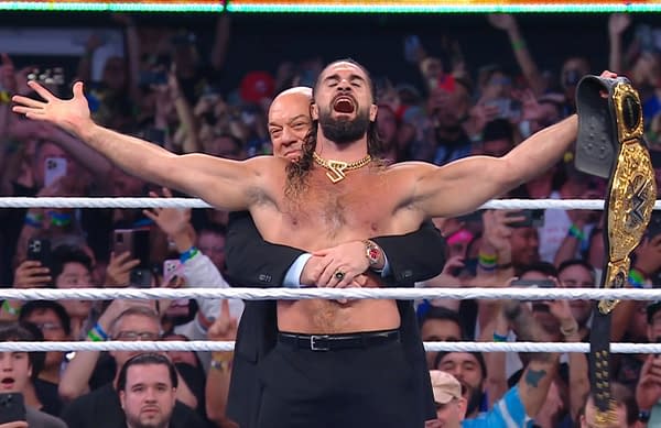 Seth Rollins celebrates in the wrestling ring, holding a championship belt high while manager Paul Heyman hugs him from behind. The crowd is cheering in the background.