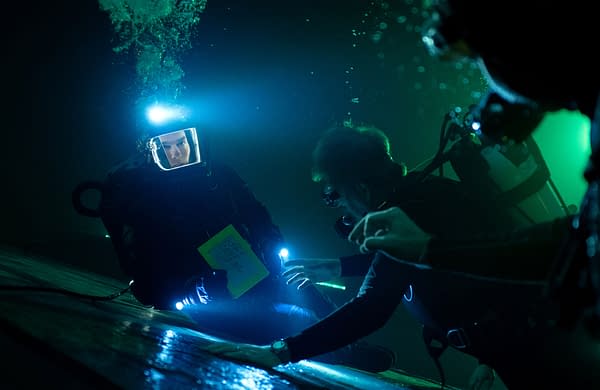 Underwater scene from 'Mission: Impossible - The Final Reckoning,' featuring a diver illuminated by bright lights, holding a yellow notepad with notes, while another diver approaches with equipment.