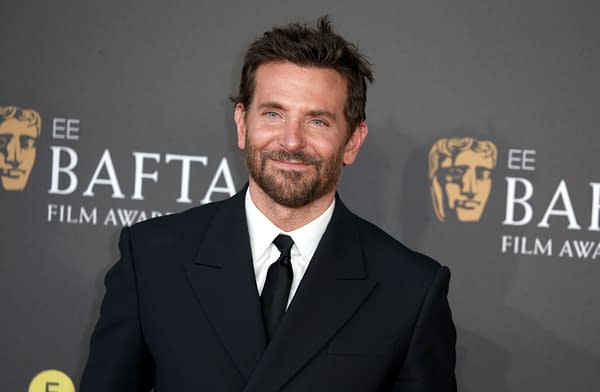 A half-length portrait of actor Bradley Cooper at the BAFTA Film Awards, wearing a black suit and tie, smiling against a dark background featuring the BAFTA logo.