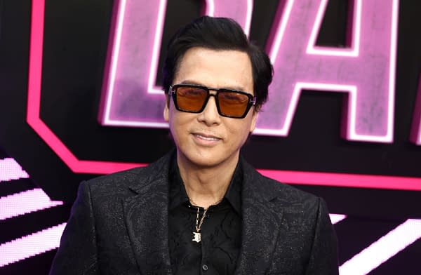 Donnie Yen poses at a film event, wearing sunglasses and a dark patterned suit against a neon pink backdrop.