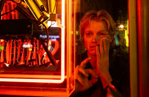A red-lit close-up of a person with a worried expression, pressing a hand to their face and looking through glass in a neon-lit setting. The scene has a moody, horror-like feel, fitting Clayface.
