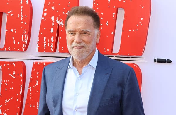 Arnold Schwarzenegger poses at a red carpet event, wearing a blue suit and light shirt, with bold red promotional signage in the background.