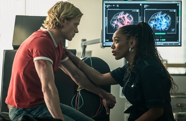 Two young adults face each other in a medical lab, with brain scan monitors glowing behind them. The man in a red shirt sits while the woman steadies his arm and speaks to him.