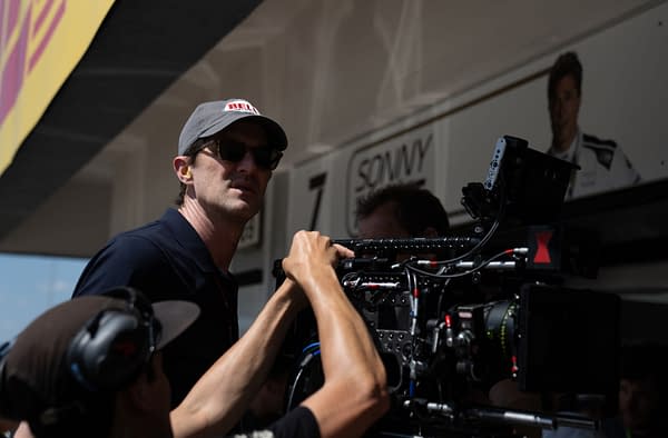 Behind-the-scenes scene at a Formula 1 event, featuring a man wearing sunglasses and a cap, overseeing camera equipment while another person adjusts it. A sponsorship banner and a wall with a recognizable name in the background provide context.