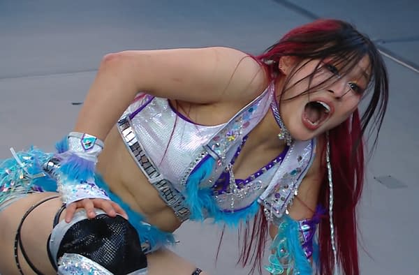 A professional wrestler with long, red-tipped hair, IYO SKY, is seen yelling passionately while wearing a sparkling, sequined outfit with blue accents. They are positioned on the mat in a wrestling ring, expressing intense emotions during a match.