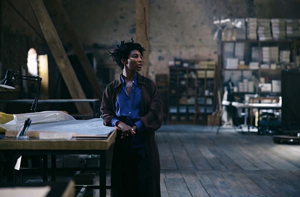 A woman stands thoughtfully in a spacious, rustic room filled with wooden beams and shelves. She wears a blue shirt and a brown cardigan, exuding a contemplative presence.